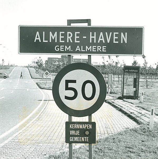 Zwart-wit foto van Stadsdeelbord Almere Haven met daaronder bordje met de tekst Kernwapenvrije Gemeente, foto Jos Jongerius uit de jaren tachtig.