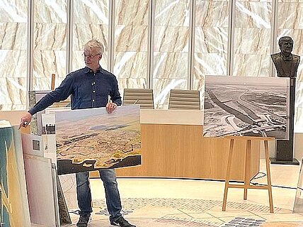 Michel Langendijk staat met een grote luchtfoto van Almere (in kleur) in zijn hand. Rechts van hem staat een andere luchtfoto in zwart-wit.